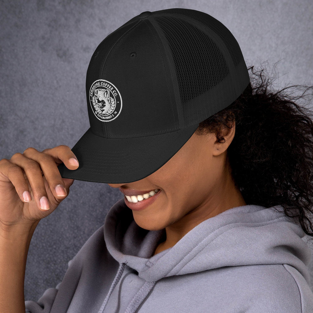 A person wearing a black trucker cap with mesh back and embroidered logo, smiling against a textured background.