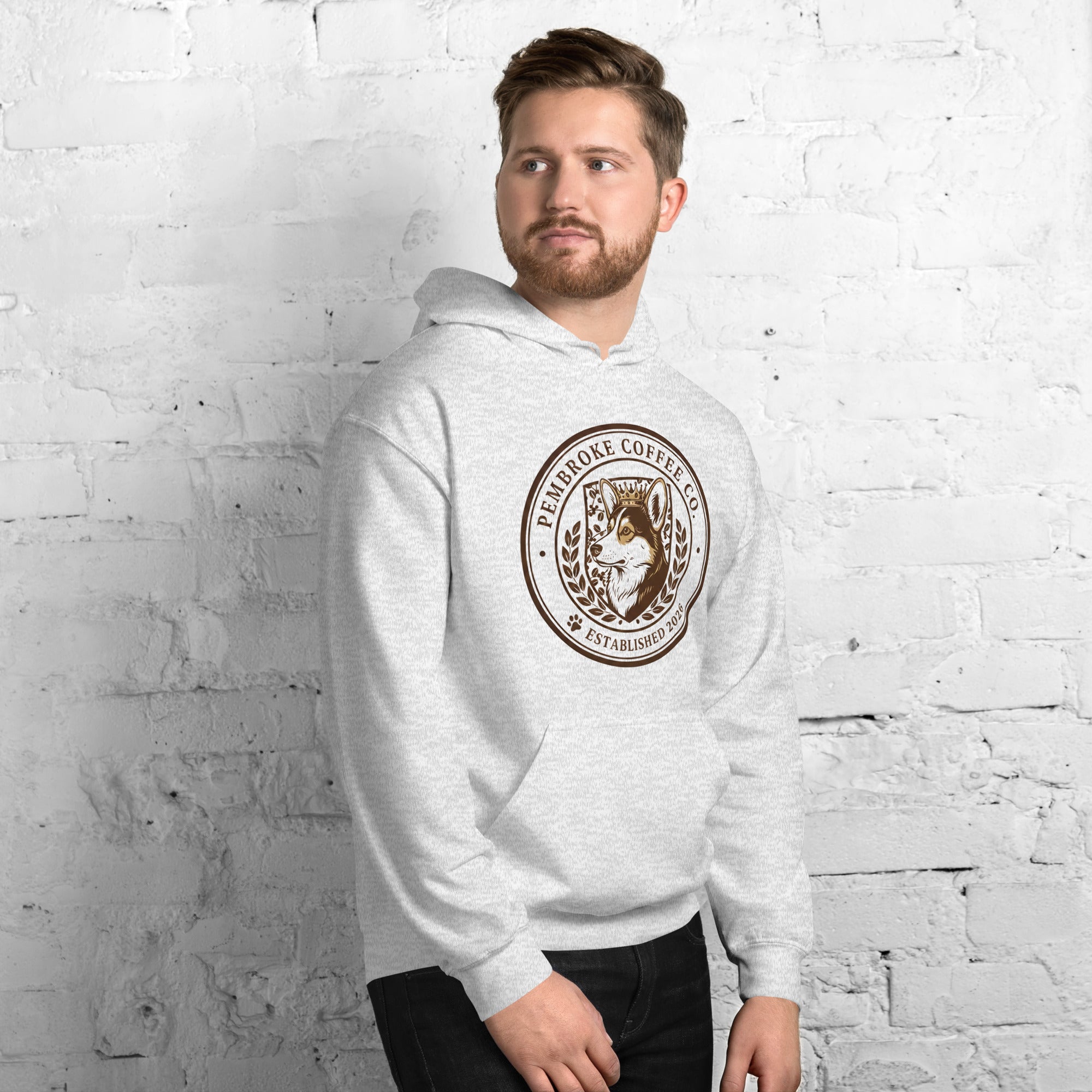 Man wearing a Unisex Hoodie featuring a logo from Pembroke Coffee Co on a brick wall background.