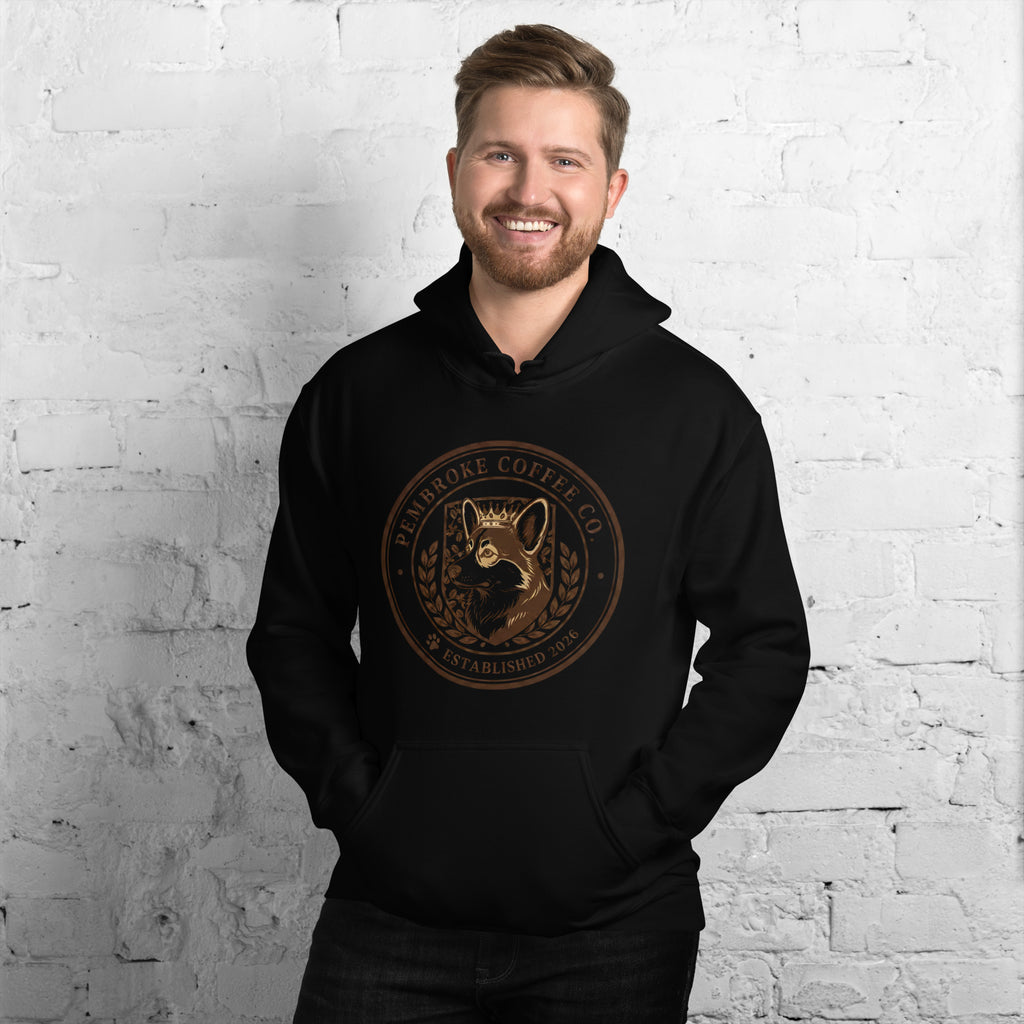 Smiling man wearing a black Unisex Hoodie featuring Eldenbrooke Coffee Co. logo against a brick wall backdrop.