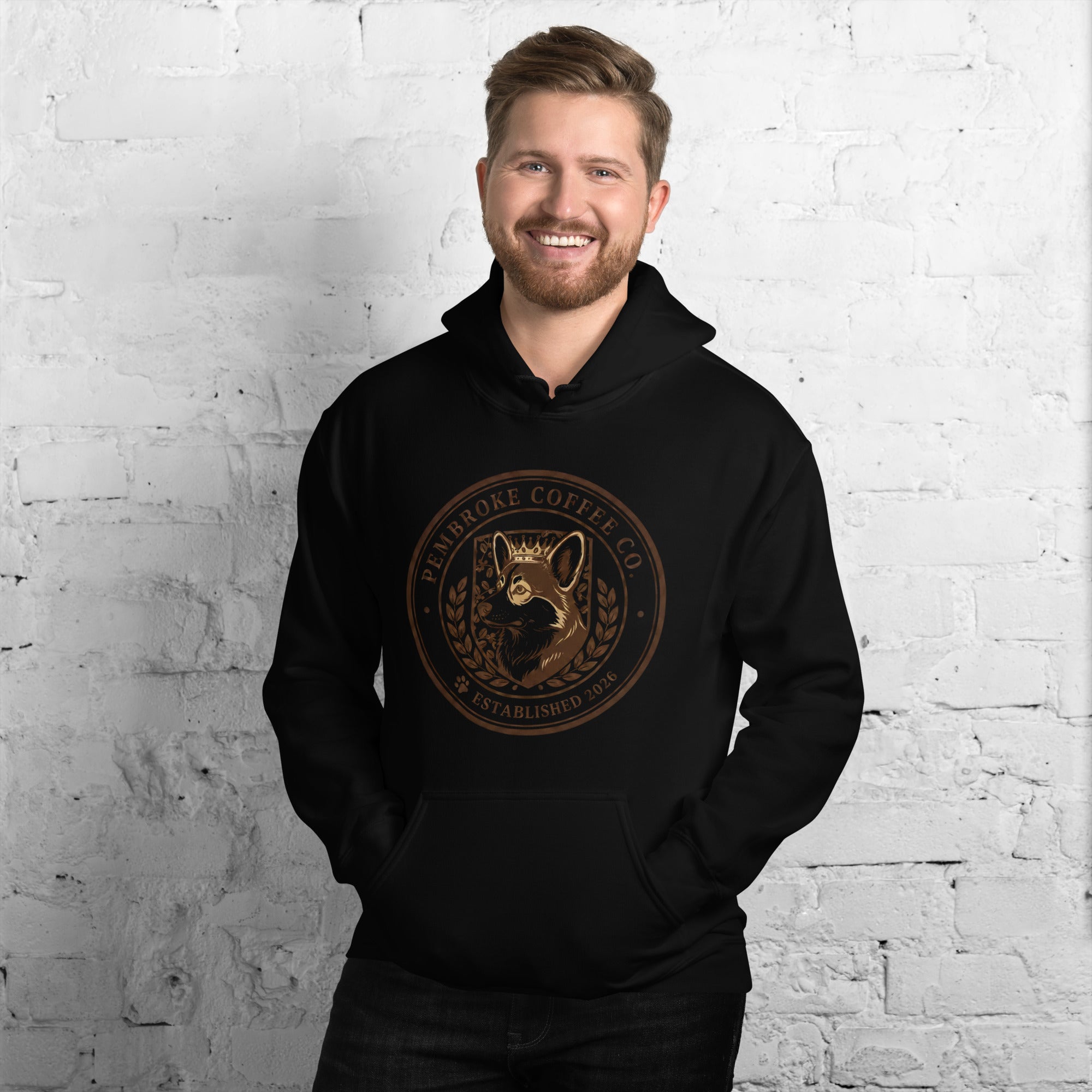Smiling man wearing a black Unisex Hoodie featuring Eldenbrooke Coffee Co. logo against a brick wall backdrop.
