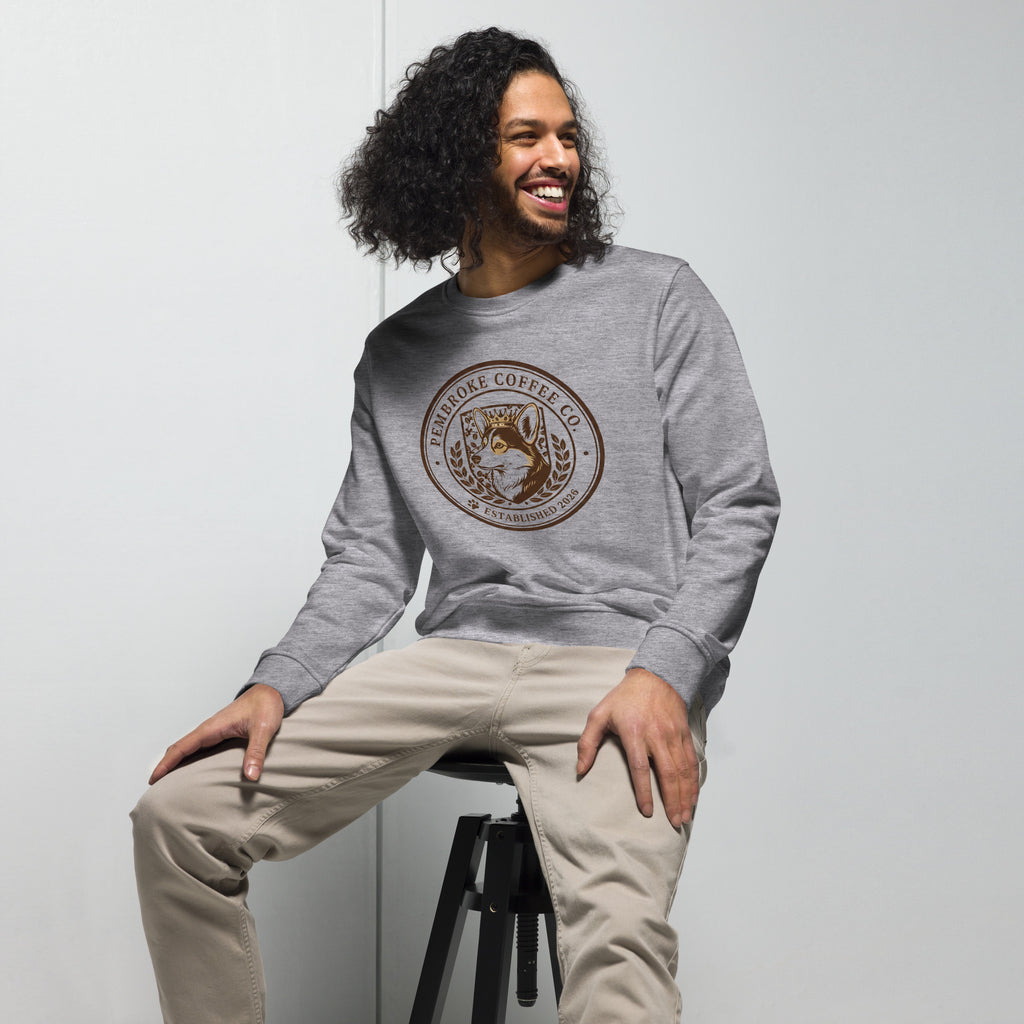 Unisex organic sweatshirt featuring a logo design, worn by a man with curly hair, sitting on a stool.